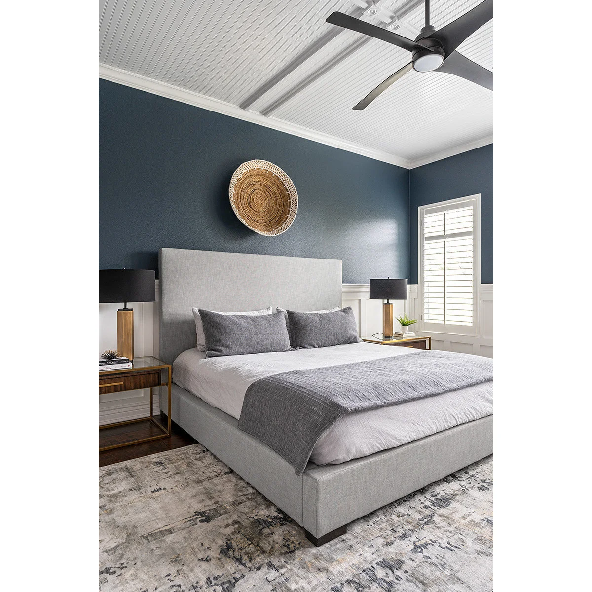 A gray bed beneath dark blue walls and white paneling, with two slim wood-and-metal nightstands and black-shaded lamps.