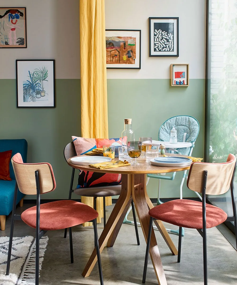 Small mixed gallery above cream and sage wall paint, with a round wooden table, coral-pink chairs, blue bench, yellow curtains, and a teal chair.