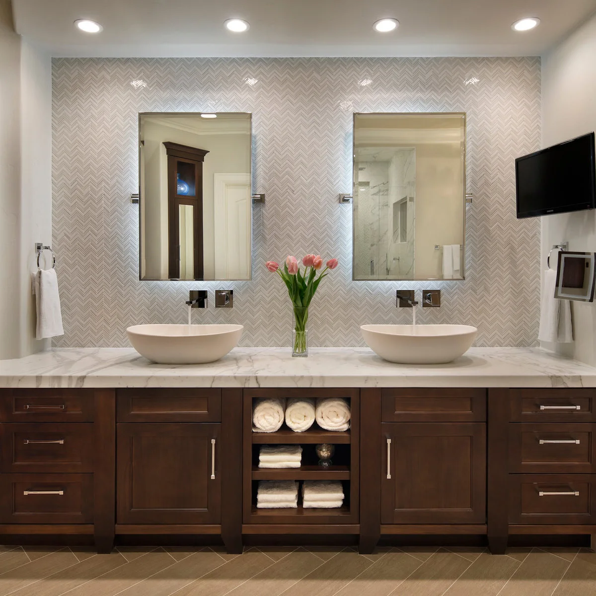 Two white oval vessel sinks sit on a pale veined counter above a dark wood vanity with open towel shelves in the center.
