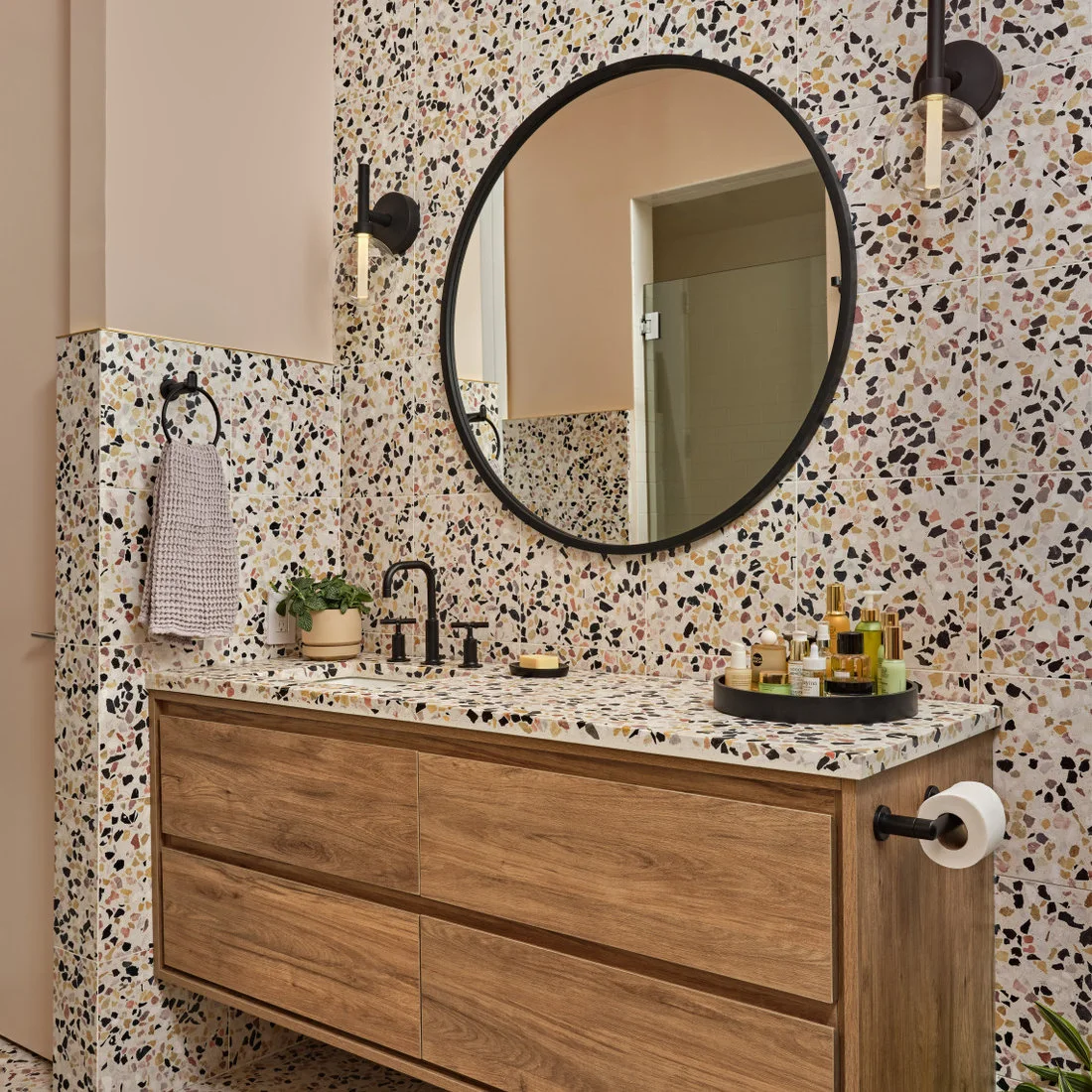 Multicolored terrazzo surrounds a timber vanity beneath a round black mirror and pale pink upper walls.