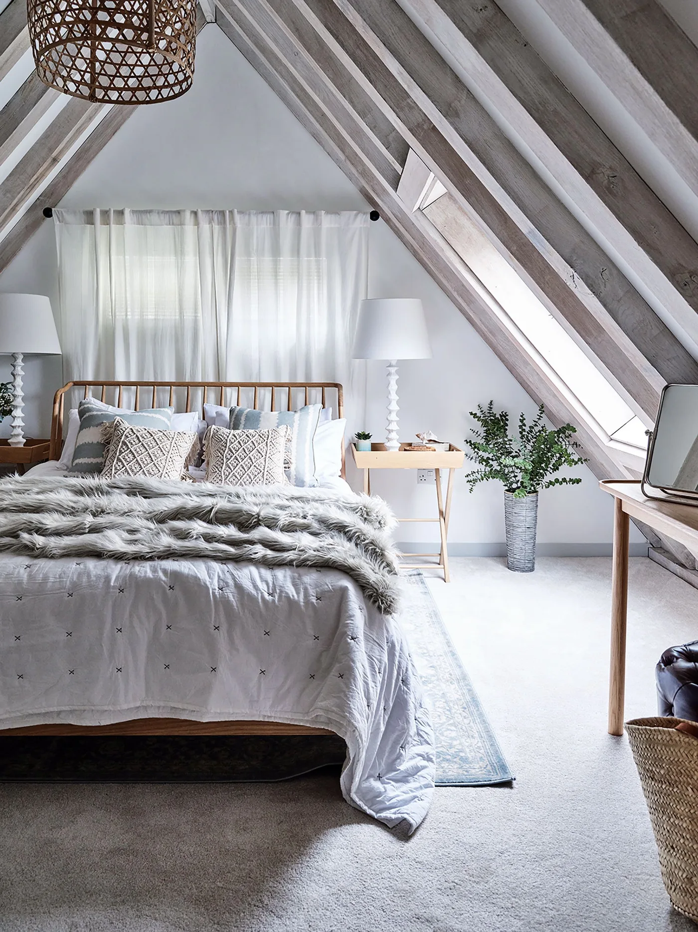 Attic bedroom with angled gray roof timbers, a wooden bed frame, pale layered bedding, two white bedside lamps and a narrow skylight.