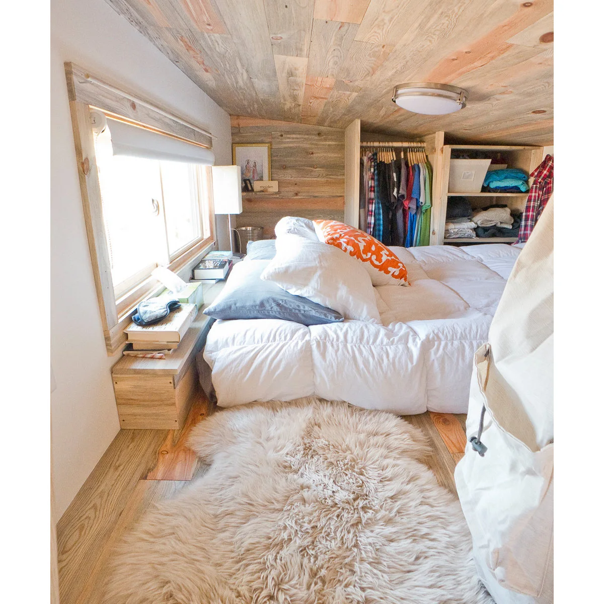 Tiny wood-lined bedroom with open clothes storage behind a bed and a narrow bedside ledge below a window