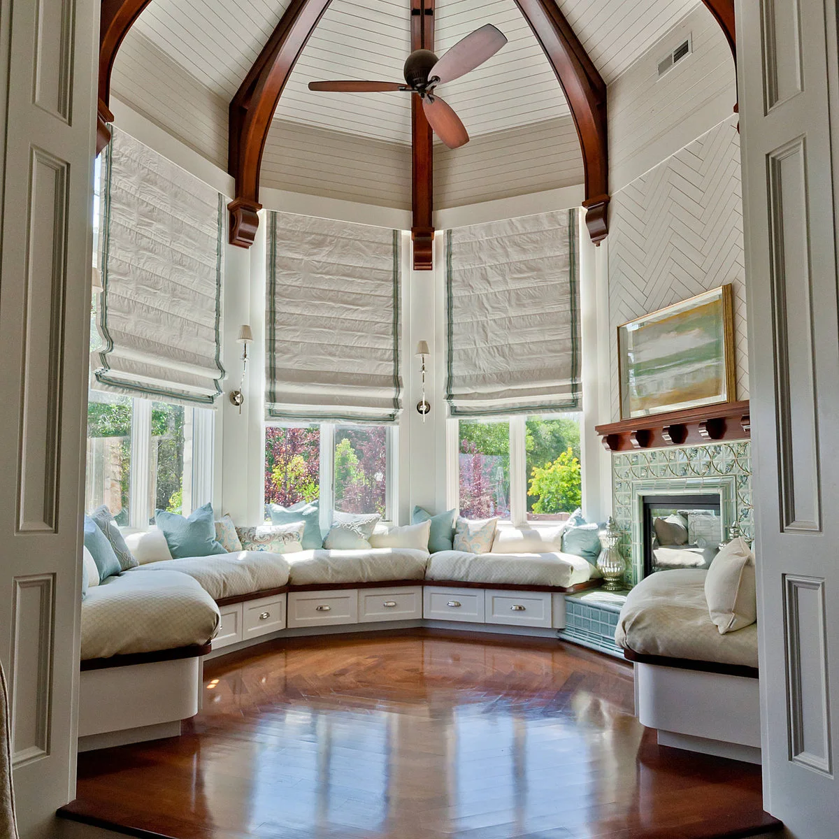A polygonal sunroom with wraparound cushioned benches, small drawers, tall Roman blinds and a tiled fireplace.