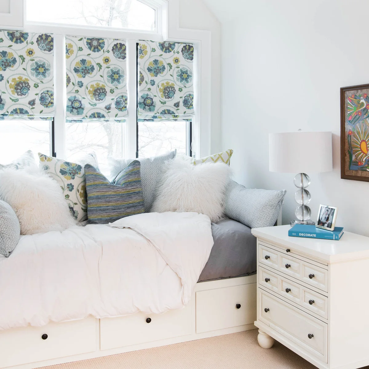 A white daybed with lower drawers has blue and green cushions that echo floral Roman shades above it.