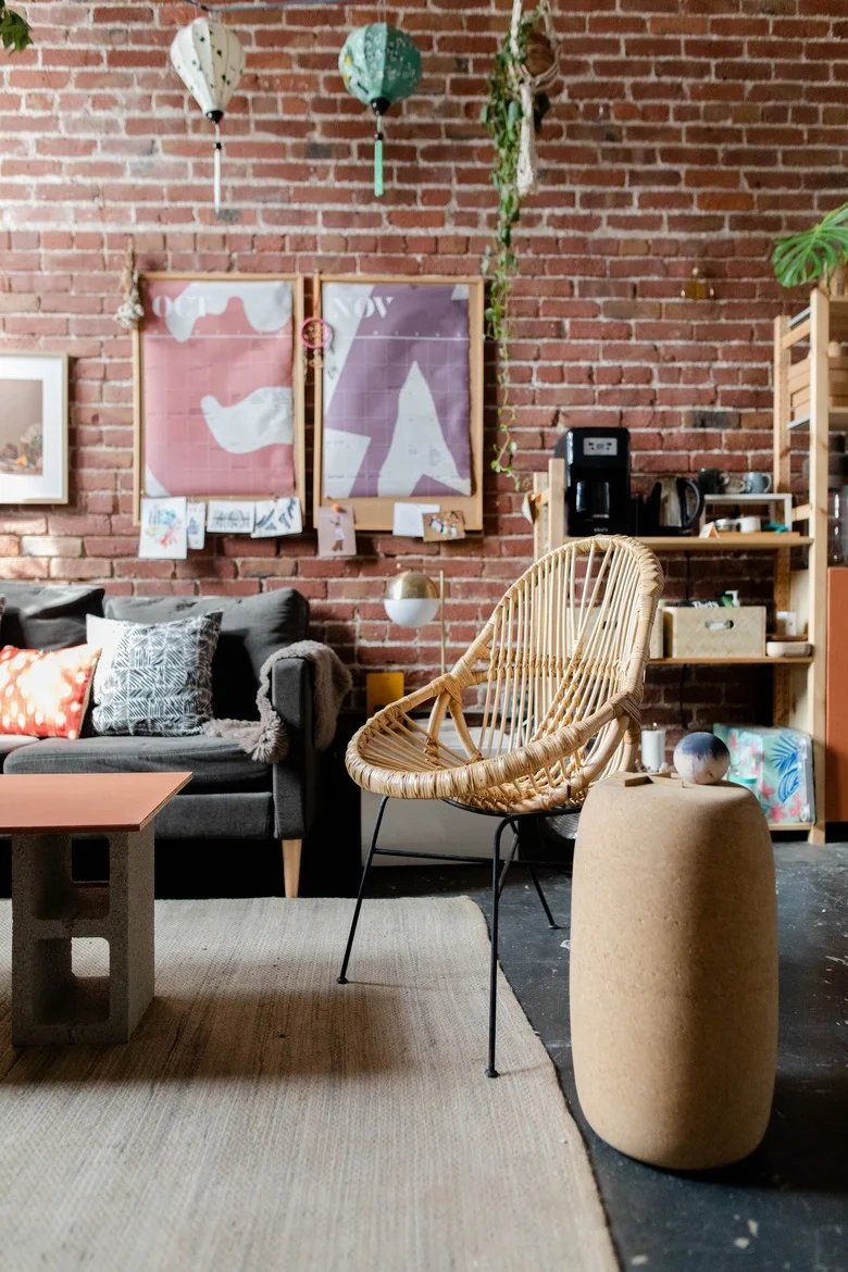A gray sofa and woven chair sit beside a brick wall, pale wood shelving, a small orange-topped table and two softly colored framed prints.