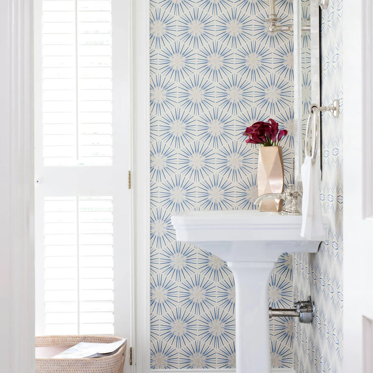 A white pedestal sink stands against blue starburst-patterned walls beside a tall shuttered window and a nearby woven container.