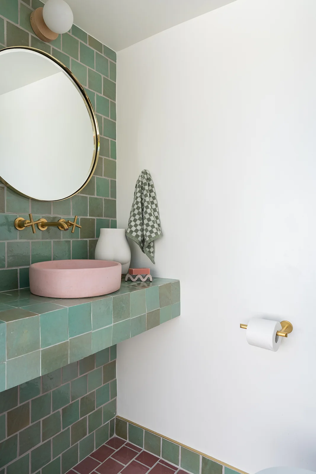A pale pink bowl basin sits on a green tiled pedestal beneath a round mirror and brass-colored wall faucet.