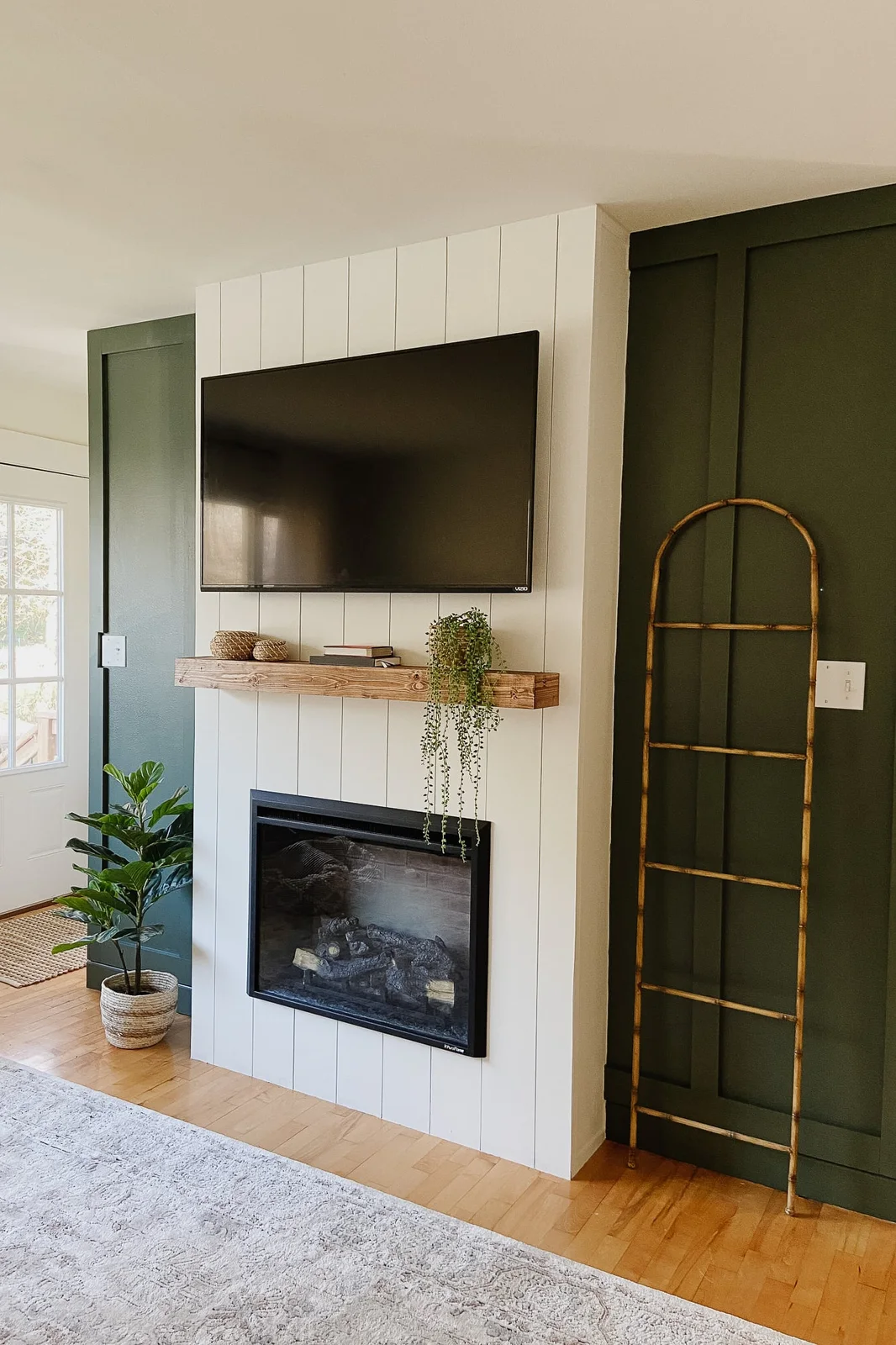 A white vertical-board fireplace wall holds a television and wood mantel between dark green paneled walls, with a trailing plant and a leaning narrow ladder.