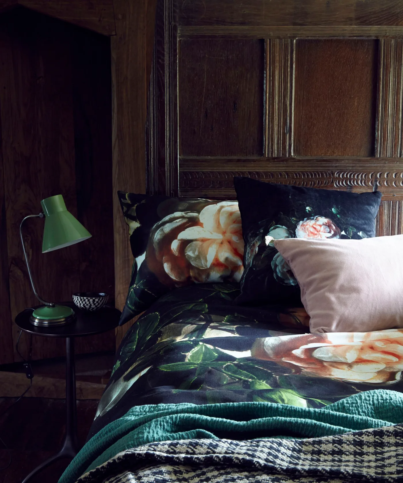 Dark carved wood bedroom paneling with large floral bedding, pink cushion, teal knitted throw, and a green bedside lamp.