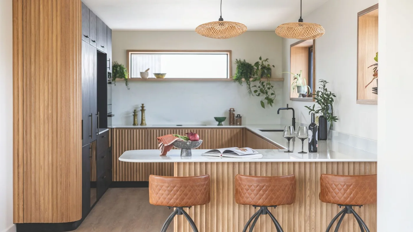A compact kitchen combines fluted oak cabinets and a peninsula with black tall cupboards, pale counters and three brown stools.