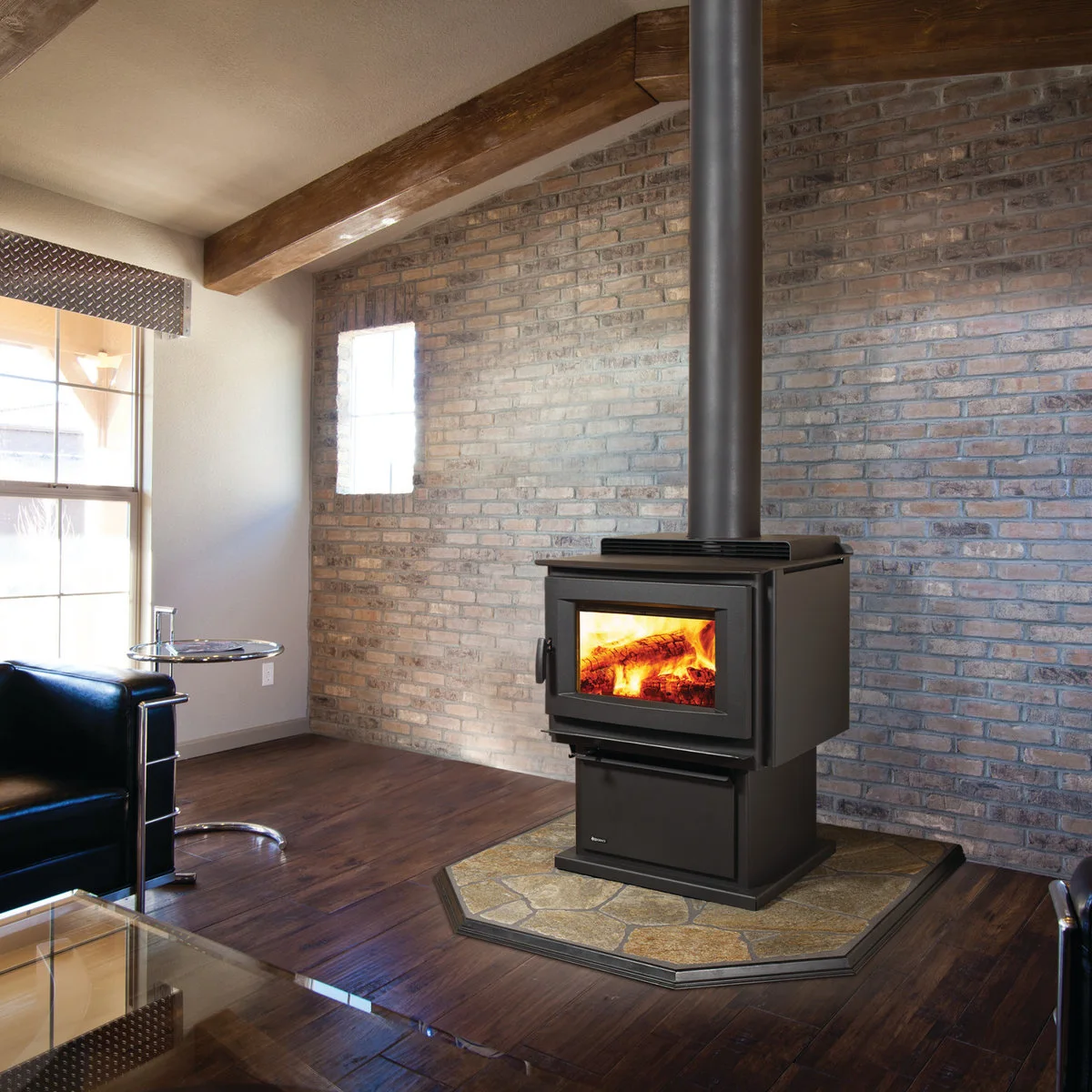 A broad black stove on a clipped-corner hearth stands against a pale varied brick wall beside dark leather seating and glass tables.