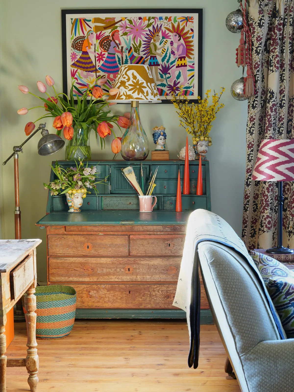 A timber chest with a green upper section sits beneath colorful framed artwork among lamps, flowers and patterned curtains.