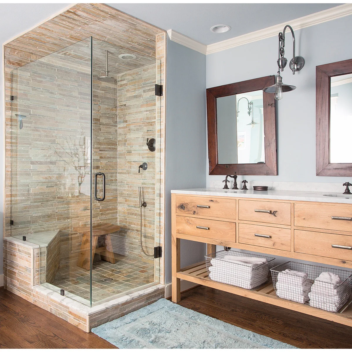 A glass-enclosed shower with slim warm-toned tiles beside a pale wood vanity, dark-framed mirrors and light blue walls.