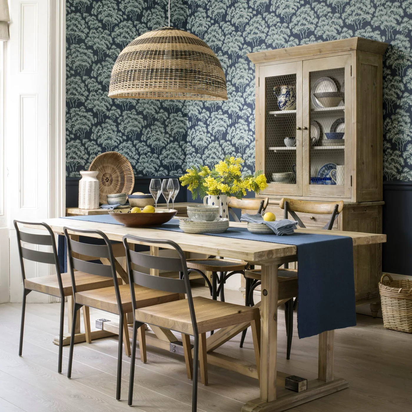 A pale timber dining table and dresser stand against navy botanical wallpaper beneath a broad woven pendant.