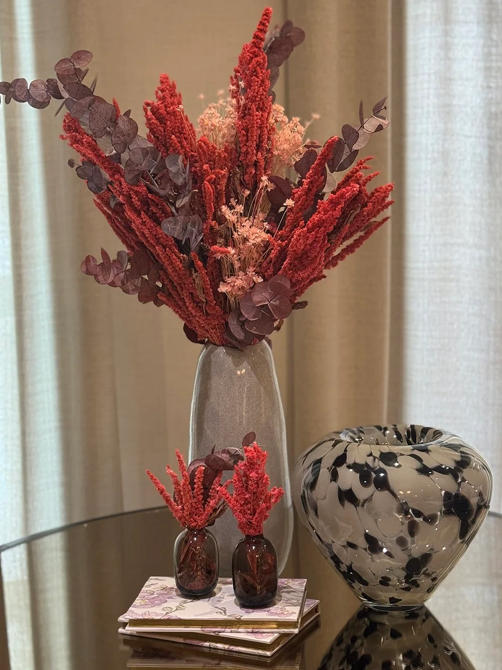 Red-toned preserved flower spikes and muted purple leaves fill a gray vase, with two small matching flower bunches in dark bud vases below.