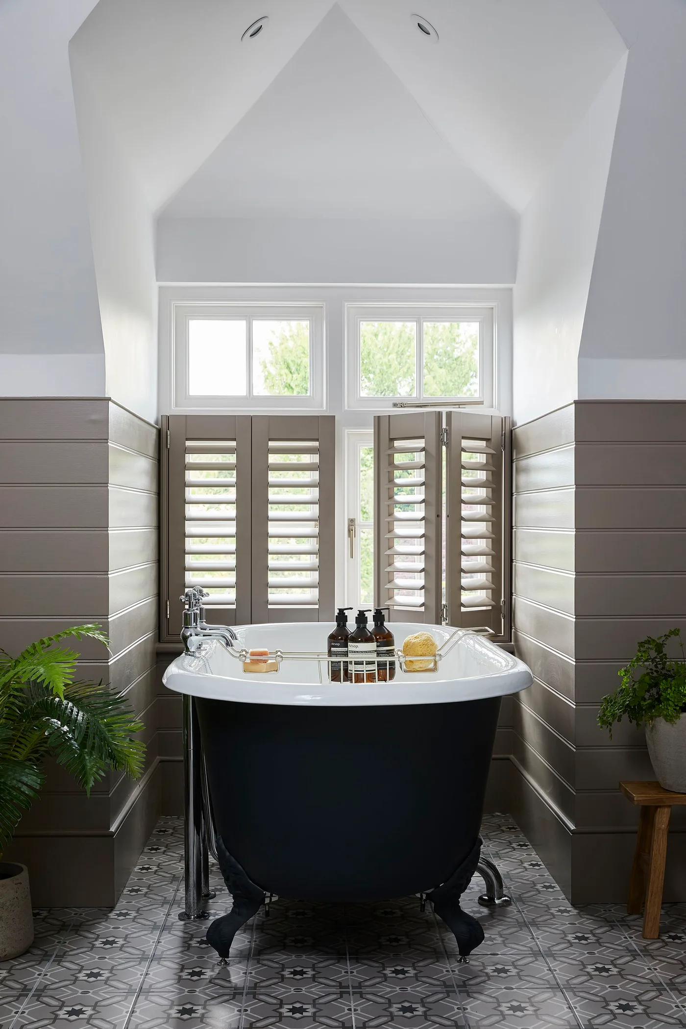 Bathroom with a dark freestanding bath, taupe horizontal paneling, lower window shutters and a patterned gray floor.