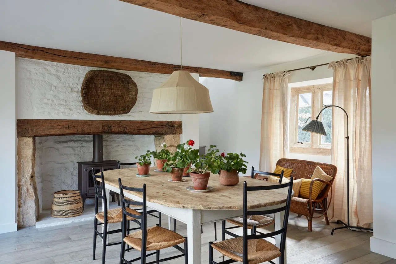 A pale weathered dining table and black woven-seat chairs stand before a white masonry fireplace beneath exposed beams.