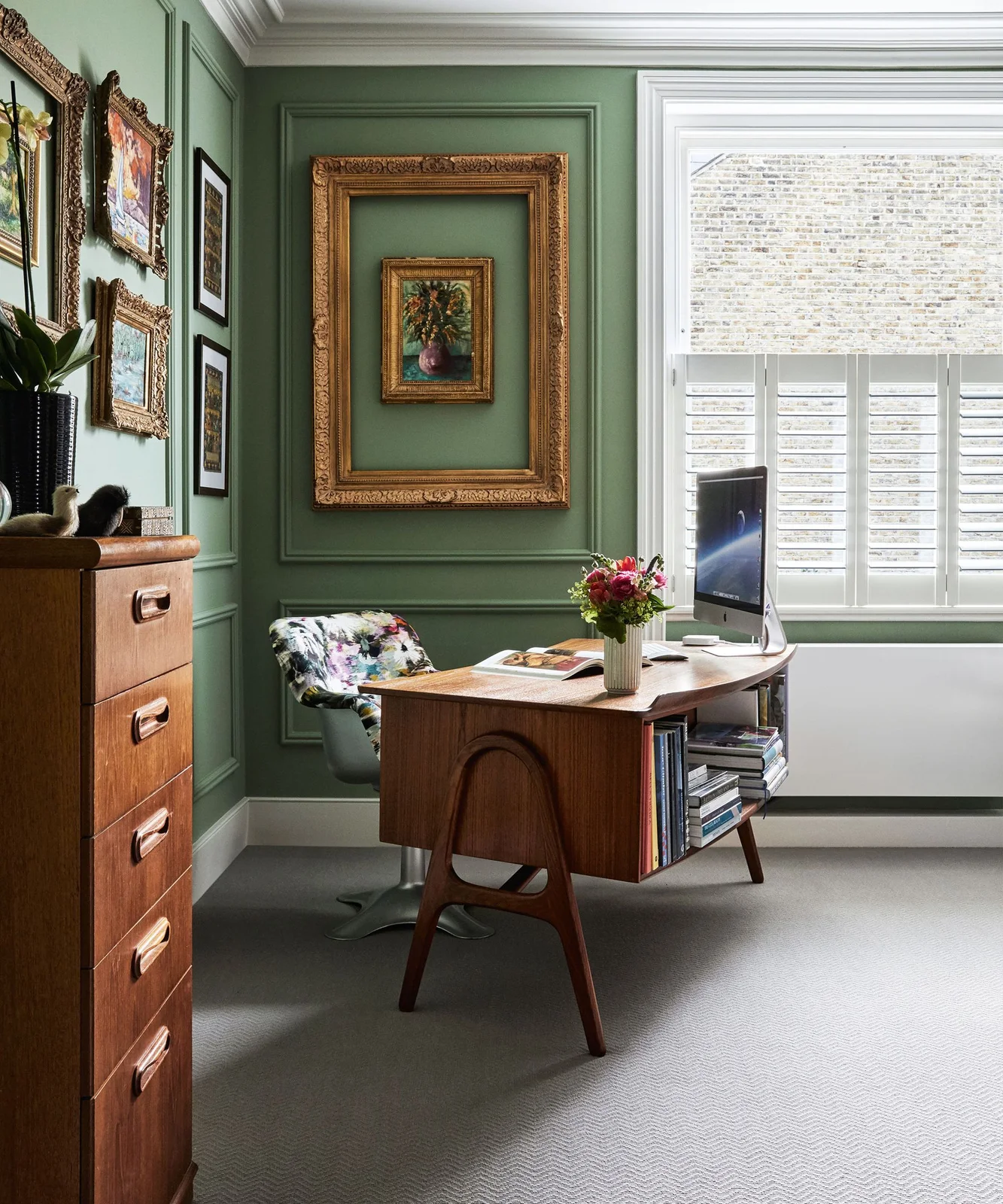 Green paneled walls with mixed framed artwork surround a rounded wooden desk beside a tall window and drawer cabinet
