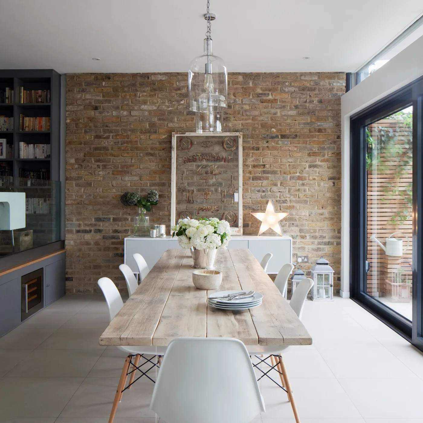 A long pale plank table with white chairs faces an exposed brick wall beneath a clear glass pendant.