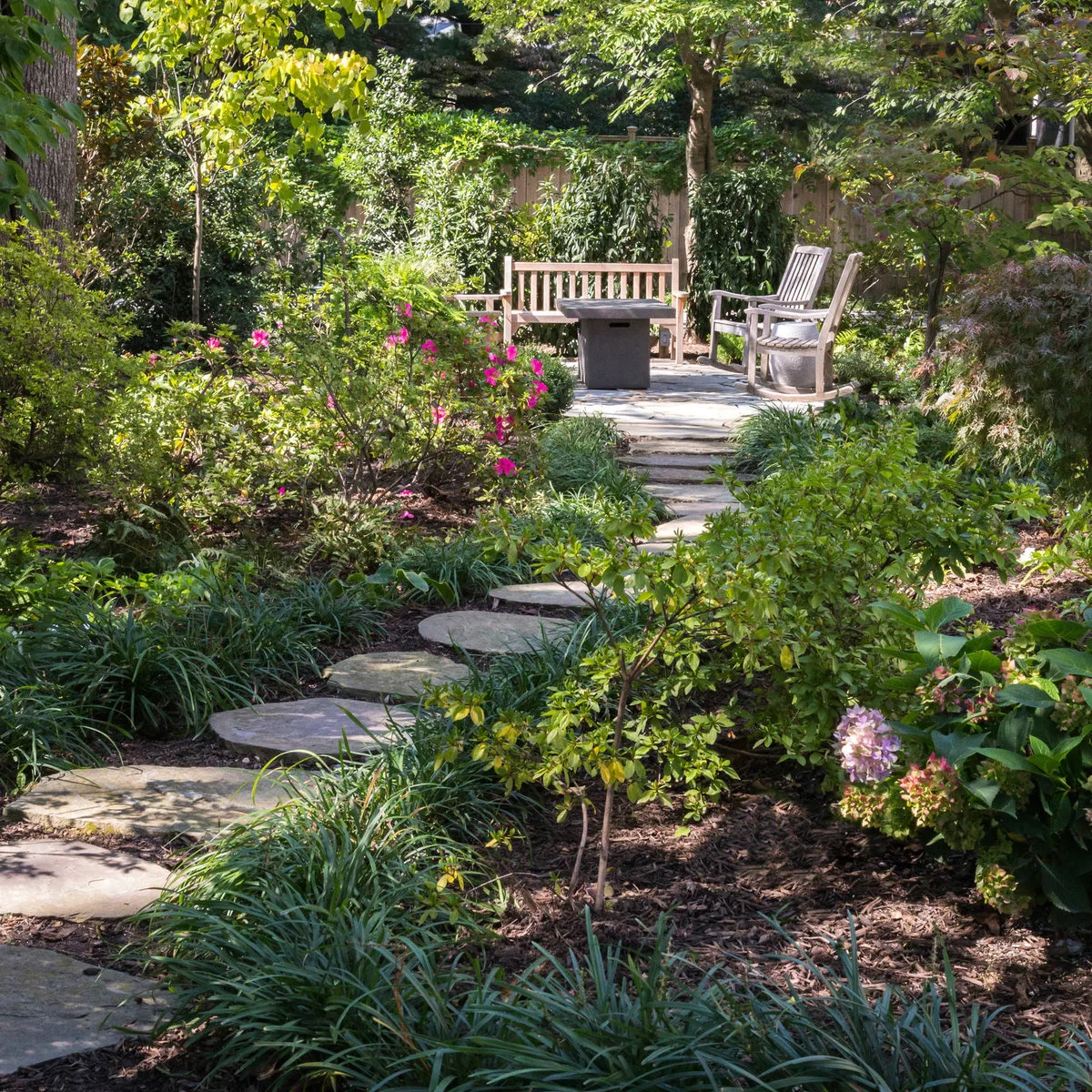 A stepping-stone path curves through dense planting toward a wood bench, two chairs and a square table.