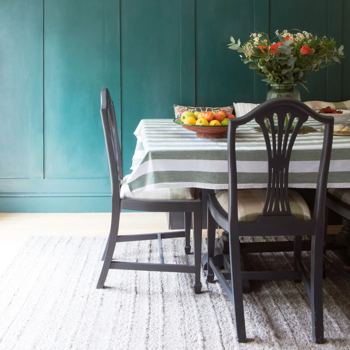 Dining room with teal wall paneling, a striped tablecloth, dark curved chairs, pale rug and flowers on the table.