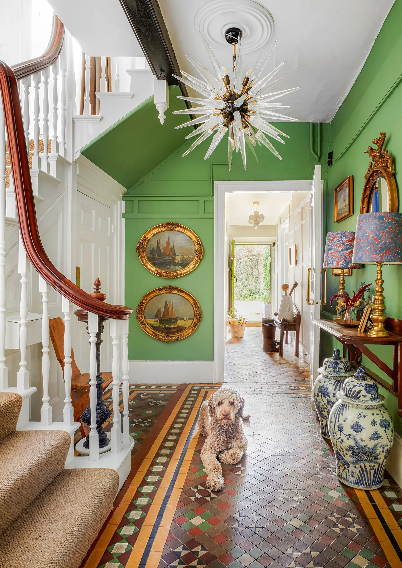 Green hallway with a white staircase, wooden handrail, ornate framed pictures and patterned floor tiles.