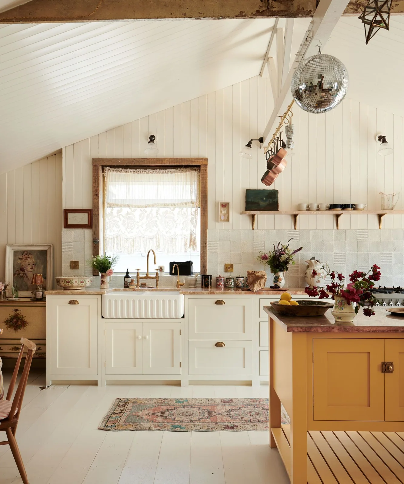 Soft ivory cabinets and a ribbed white sink sit beside a yellow island beneath pale wall boards and exposed beams.