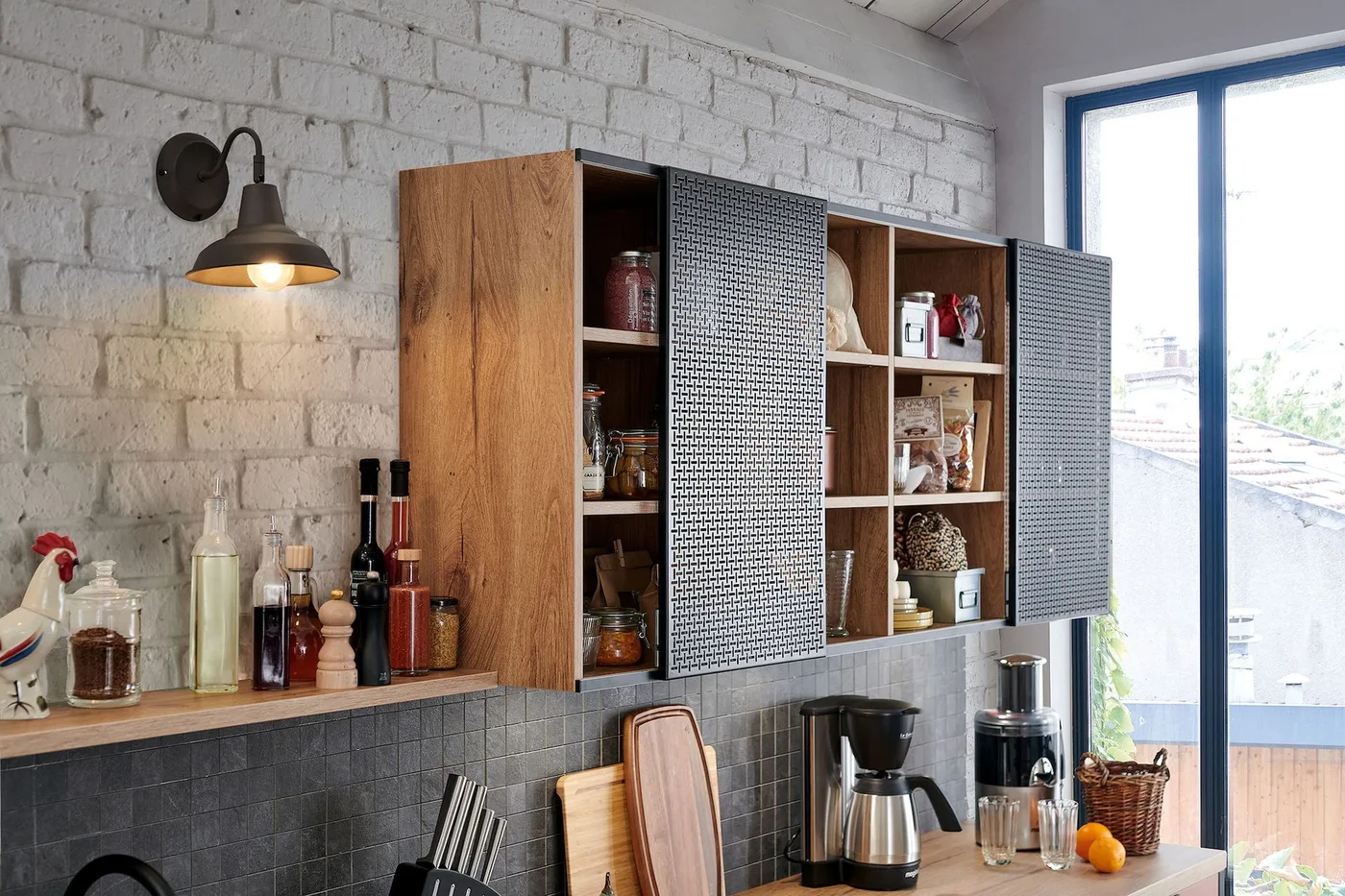 Wood-toned open cupboards with dark textured sliding fronts sit above a counter holding a coffee machine and jars beside white-painted brick and blue trim