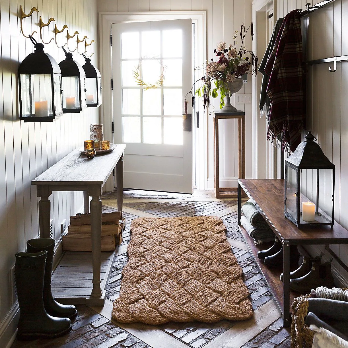 Entry mudroom with white glazed door, wood furniture, wall hooks, hanging lanterns, boots and a broad woven-pattern doormat