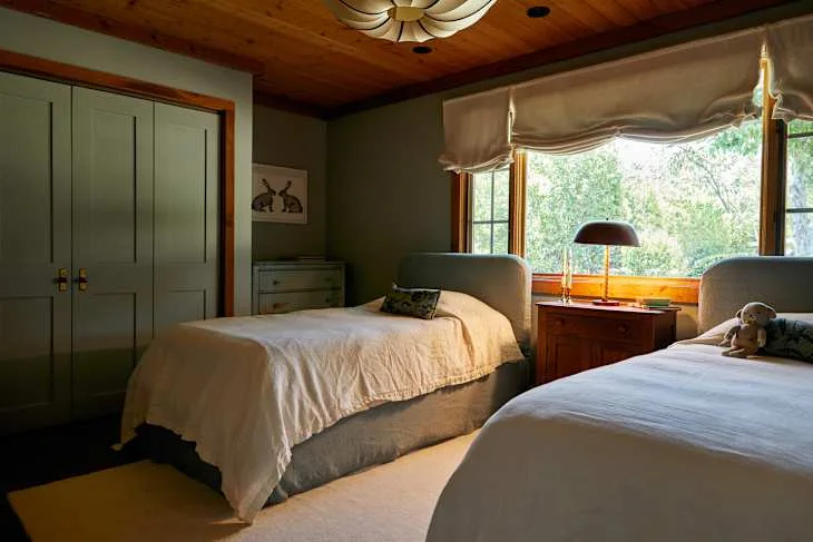 Two pale single beds stand beside green walls and matching closed wardrobes below a wood ceiling and broad shaded window