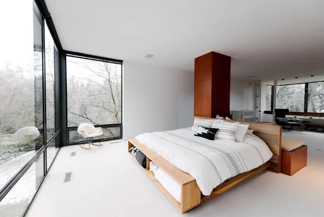 A timber platform bed sits beside floor-to-ceiling glazing in a restrained pale room.