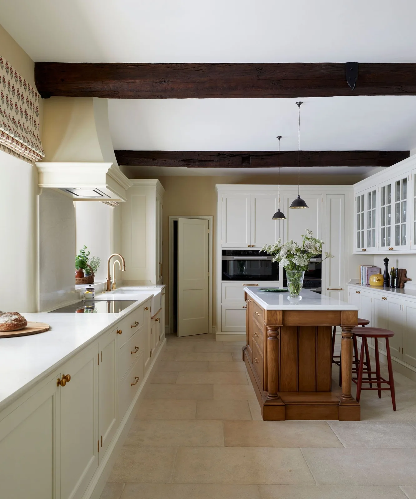 A white country kitchen has dark ceiling beams, a narrow wood island with turned posts, red stools, glazed wall cupboards and pale rectangular floor tiles.