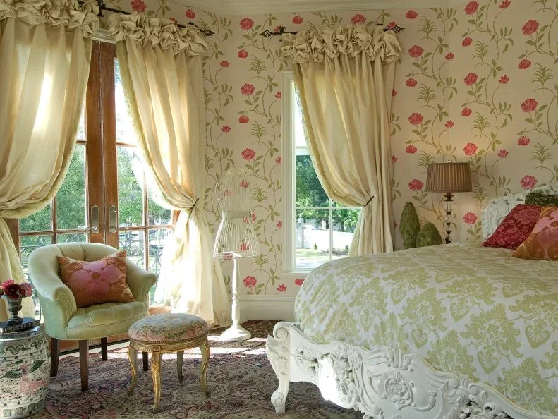 Pink rose wallpaper and green stems surround a white carved bed beside cream curtains and a pale green chair.