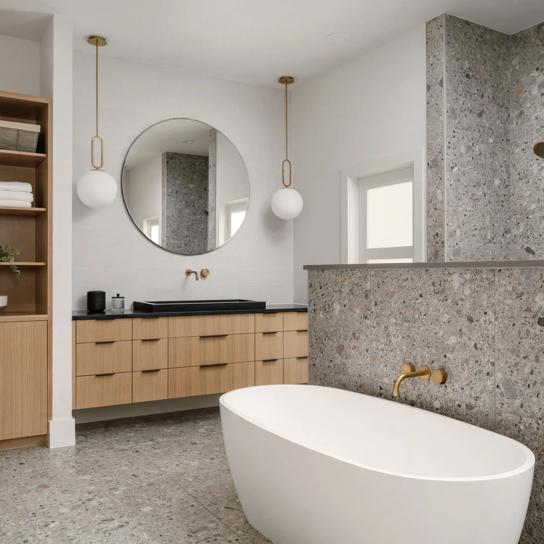 Gray terrazzo covers a partition and floor beside a white tub, black basin and timber vanity.