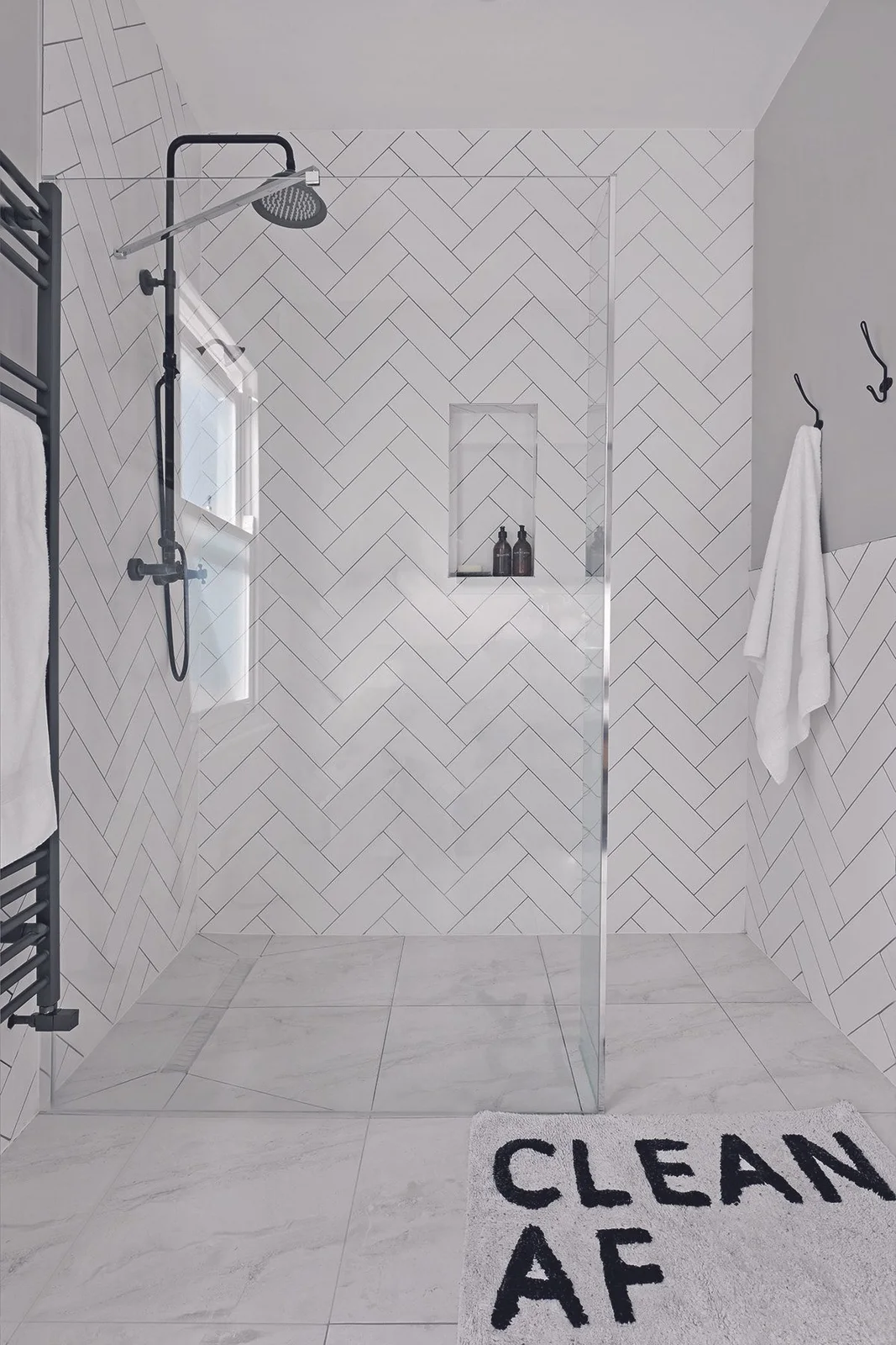 White herringbone shower walls surround a small niche beside black fittings, a clear screen, black wall hooks and pale flooring