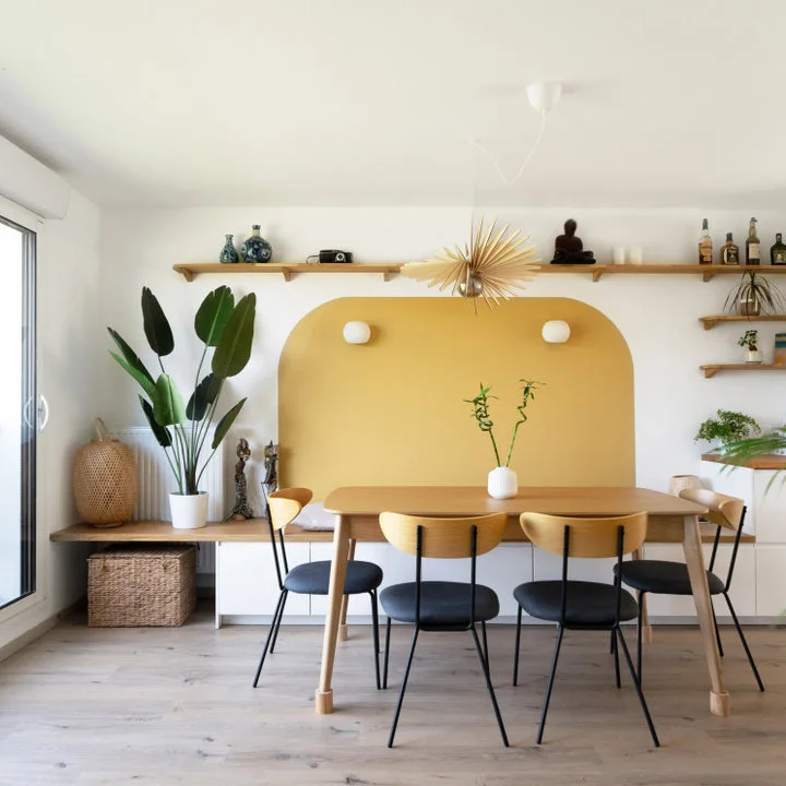 A wood dining table and dark chairs sit before a rounded mustard wall shape with a long wood display shelf above.
