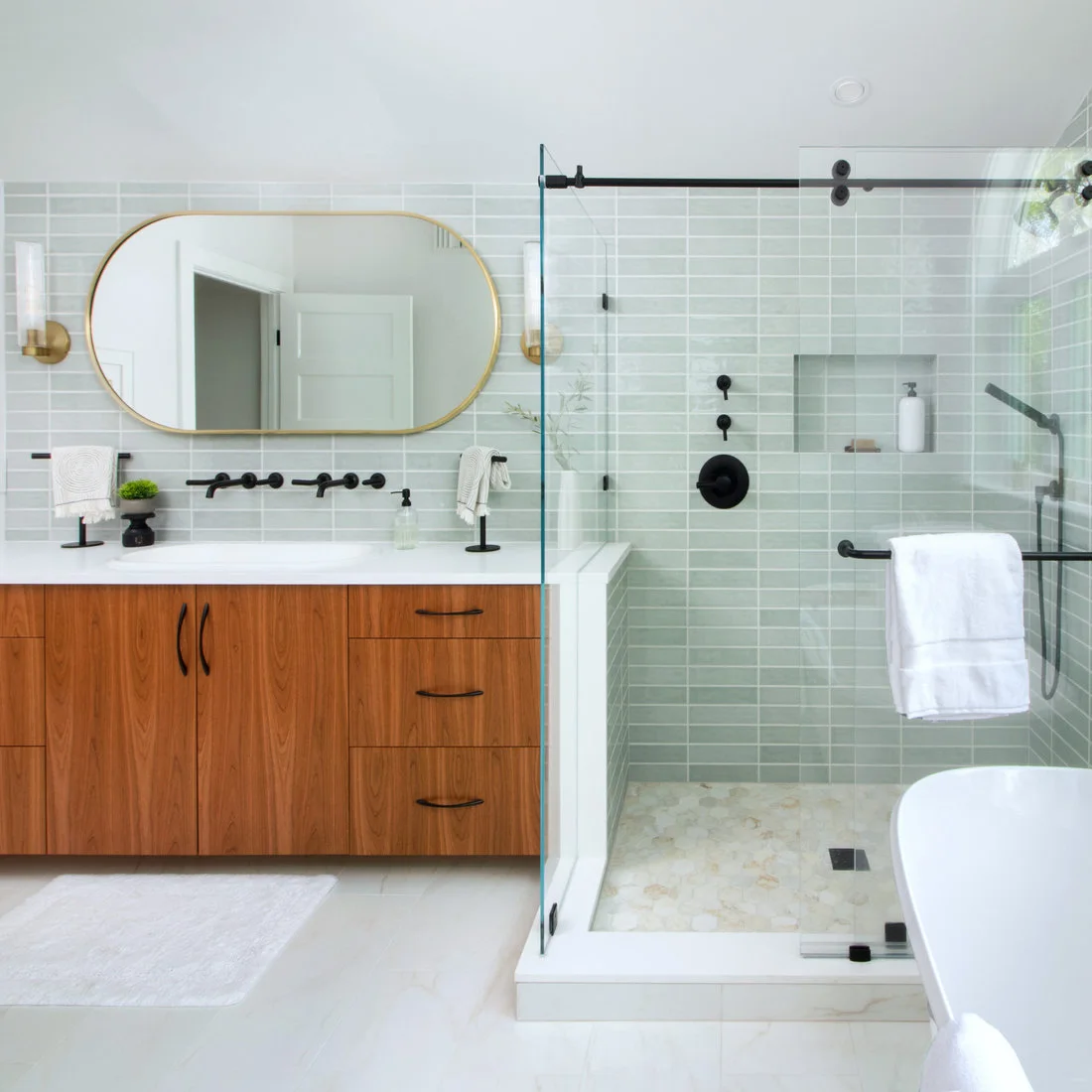Pale green rectangular tiles run behind a rounded mirror and glass shower beside a warm wood double vanity