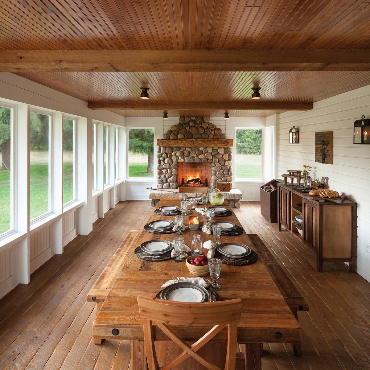 A long wooden dining table and chairs in an enclosed porch with white-framed openings, wood ceiling and stone fireplace.