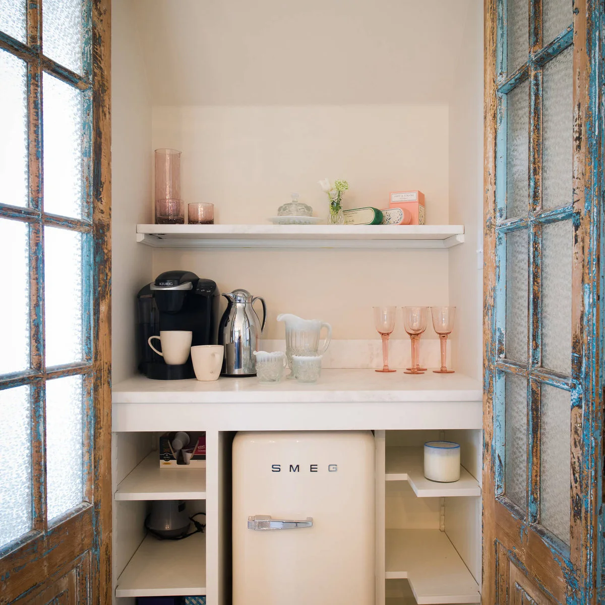 Compact drinks cupboard with distressed blue doors, coffee machine, glasses, small cream refrigerator and open shelves