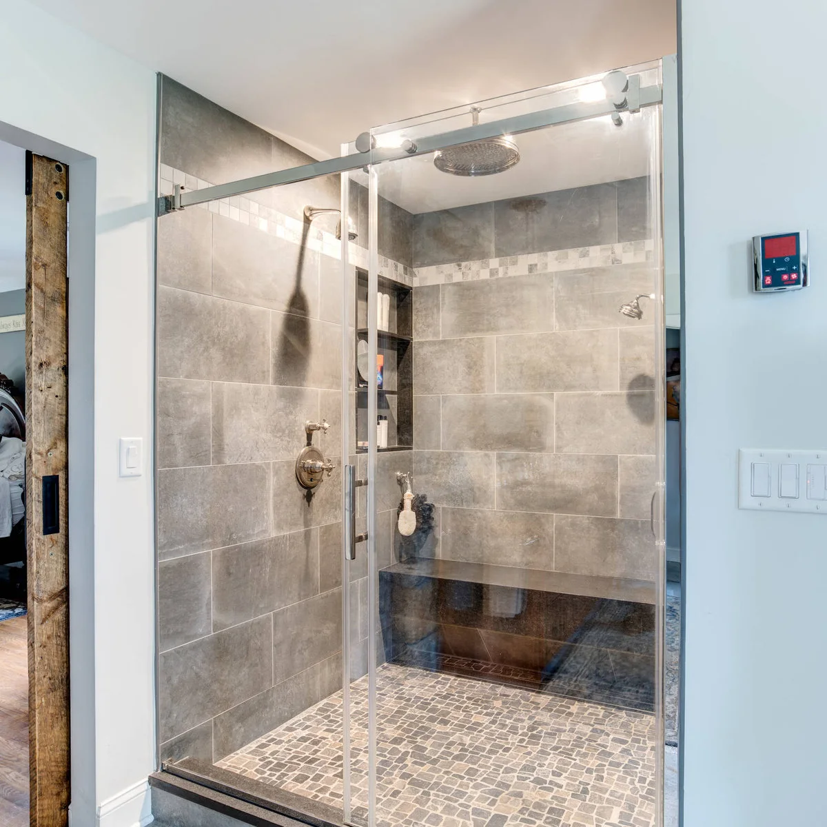 A gray-tiled shower with a sliding glass door, narrow mosaic border, recessed storage and a dark full-width bench.