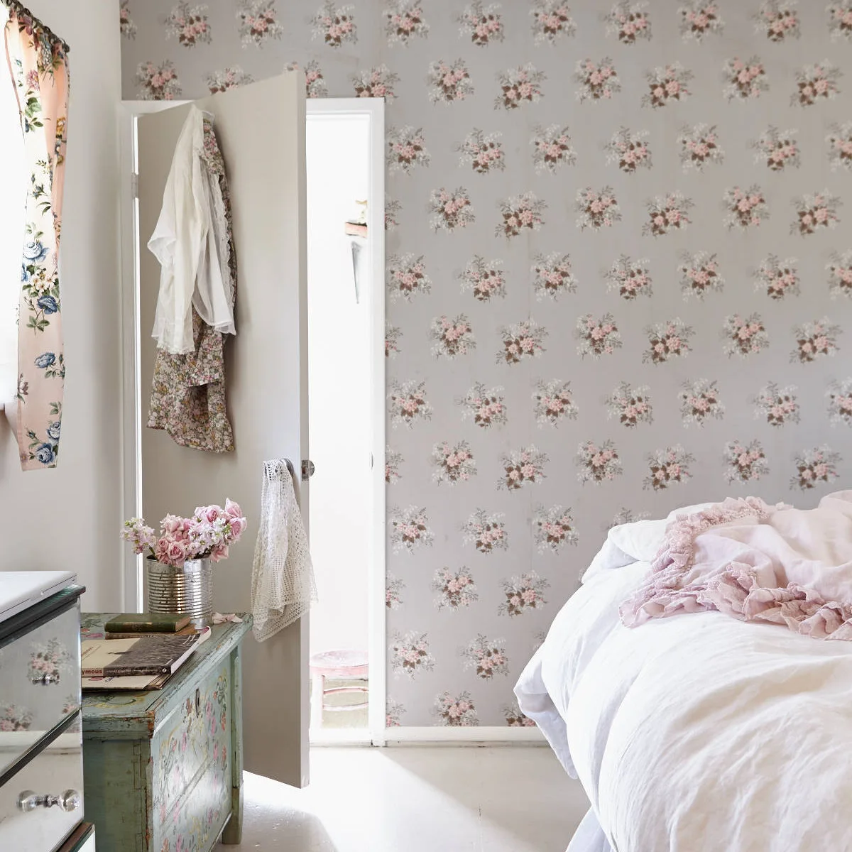 Bedroom with floral wallpaper, pale bedding and a worn painted chest beside the bed
