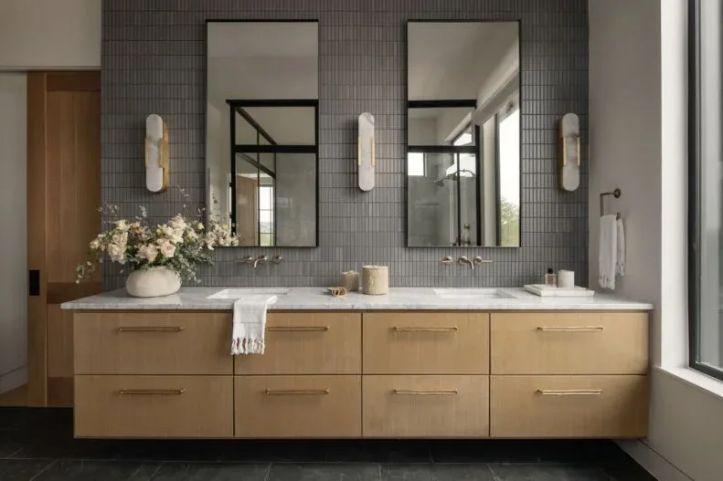 Double vanity with light wood drawers, pale counter, gray narrow vertical wall tiles, rectangular mirrors, three pale wall lights, and charcoal floor.