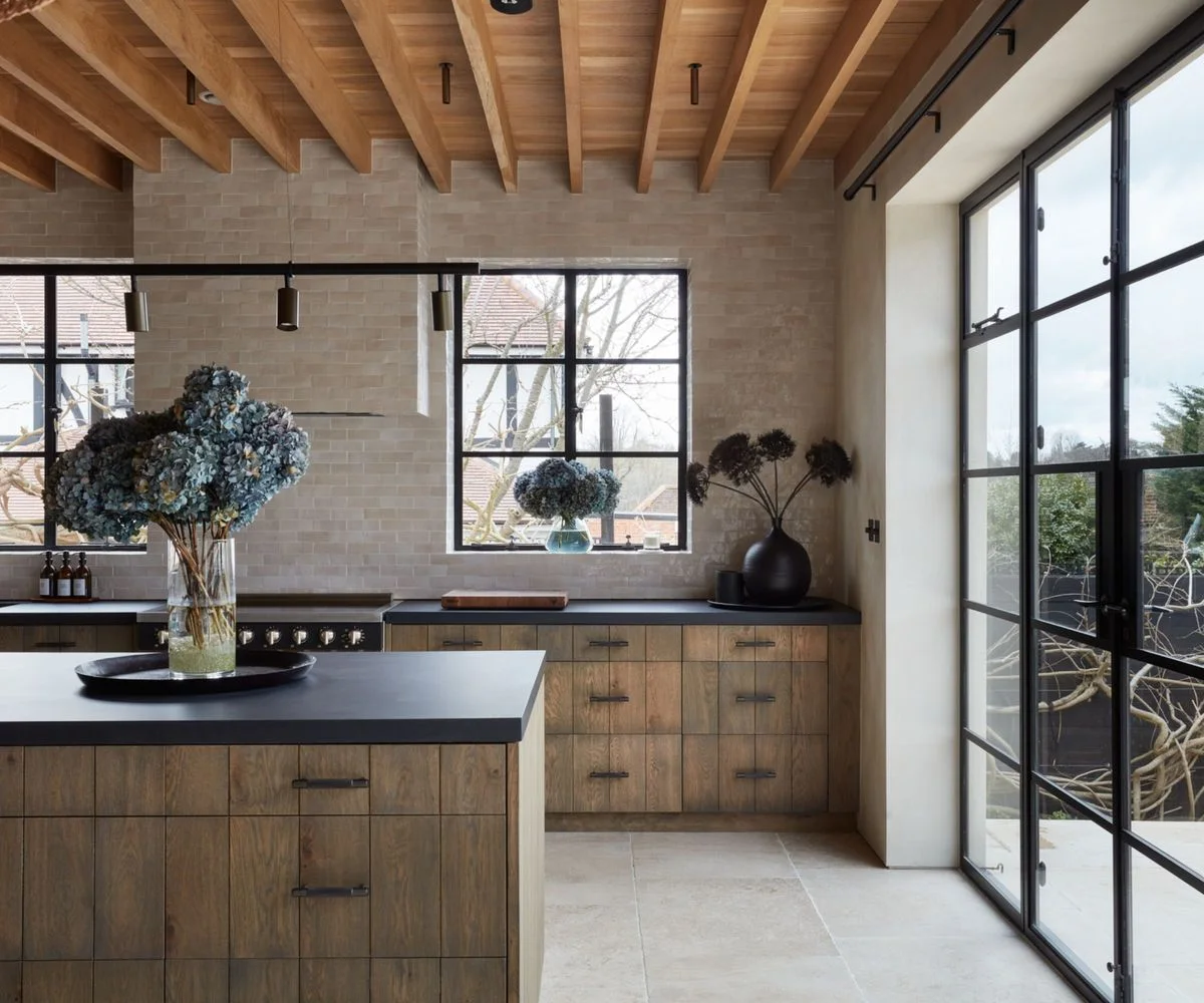 Wooden cabinets with dark counters stand beneath exposed wooden ceiling beams beside pale walls, black-framed windows and broad garden doors