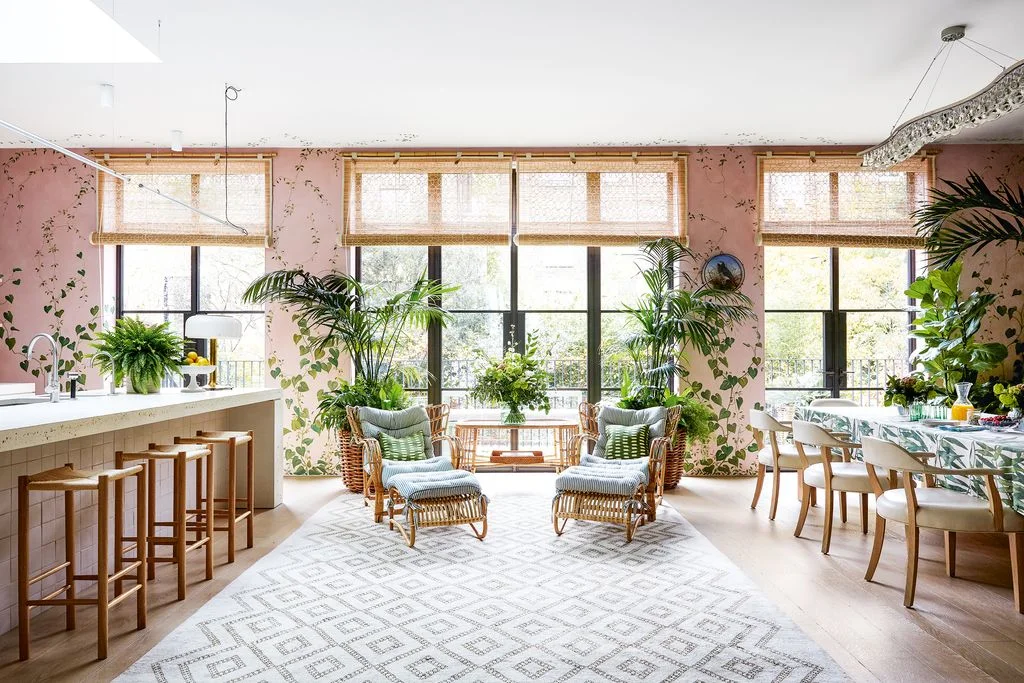 Two woven armchairs and footstools sit between a kitchen counter and dining table beside tall windows and pink botanical walls.