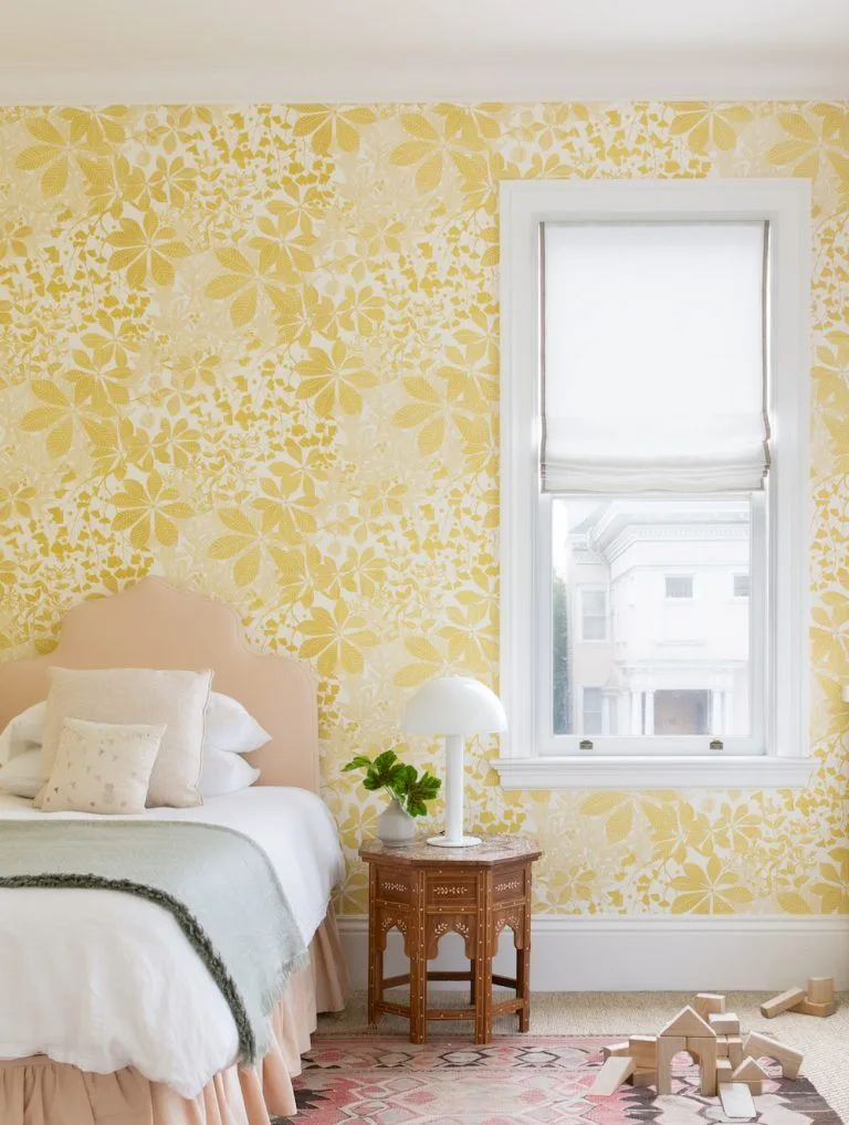 Large yellow flower wallpaper around a window beside a pale headboard and light green bedding.