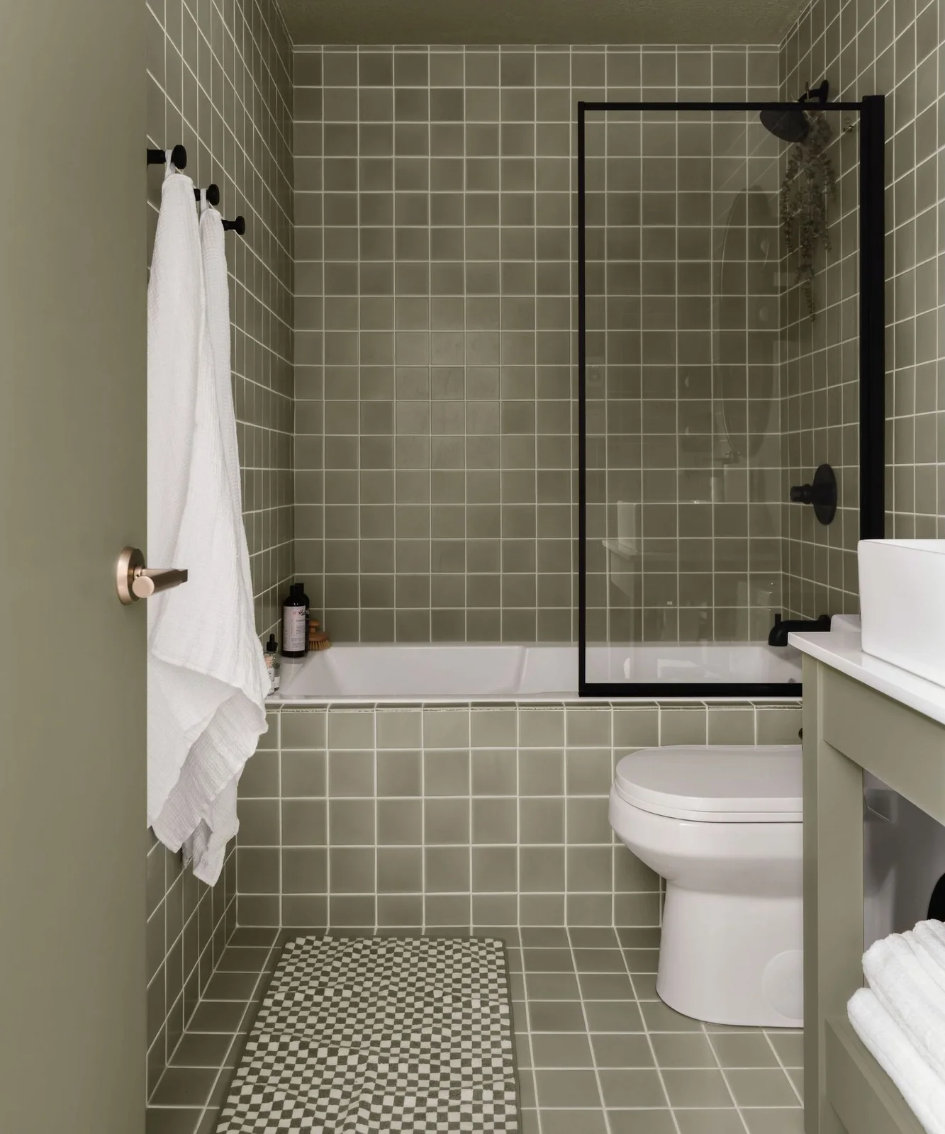 Muted green square tiles cover a tub-shower wall and tub front with pale grout, black fittings and a small checked floor