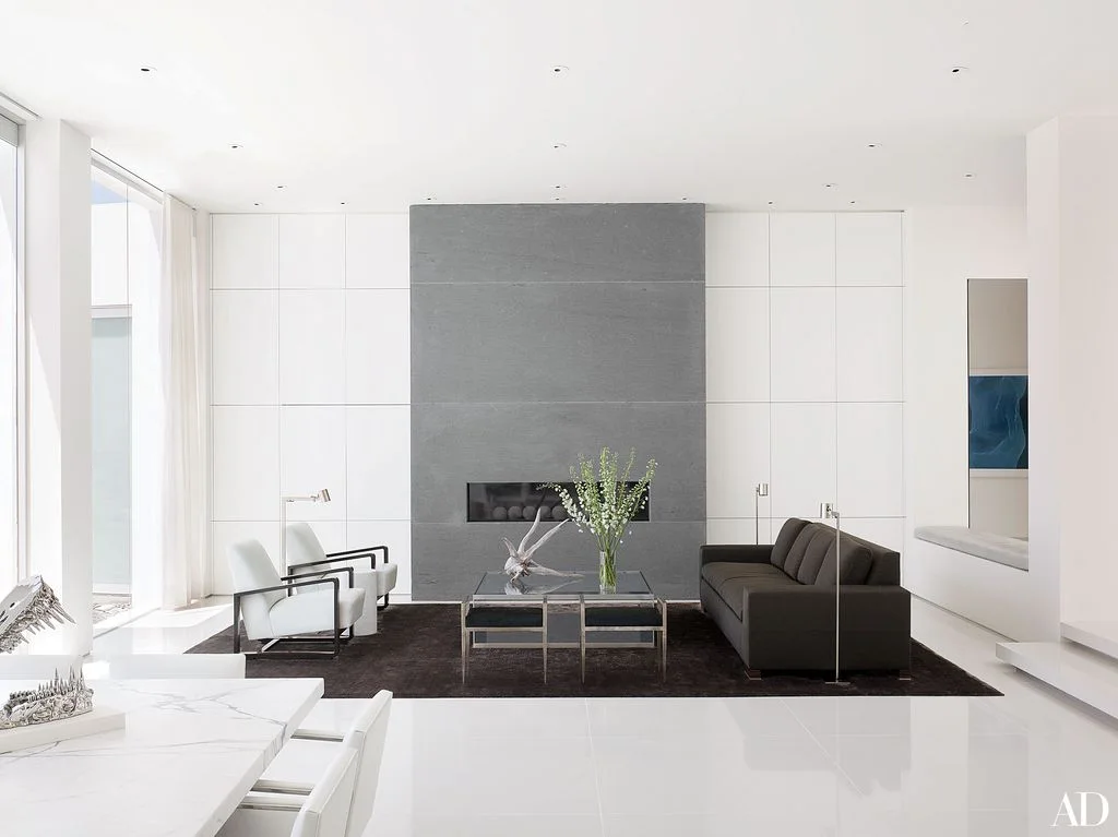 A gray central fireplace wall with a long narrow opening stands between white flat-fronted cabinets beside a dark sofa, pale chairs and a glass coffee table