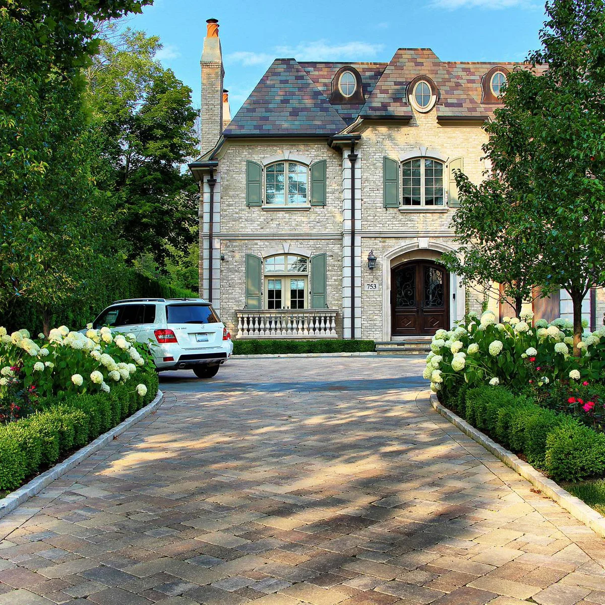 Pale curb stones and clipped low hedges frame white flowering shrubs along both sides of a paved driveway.