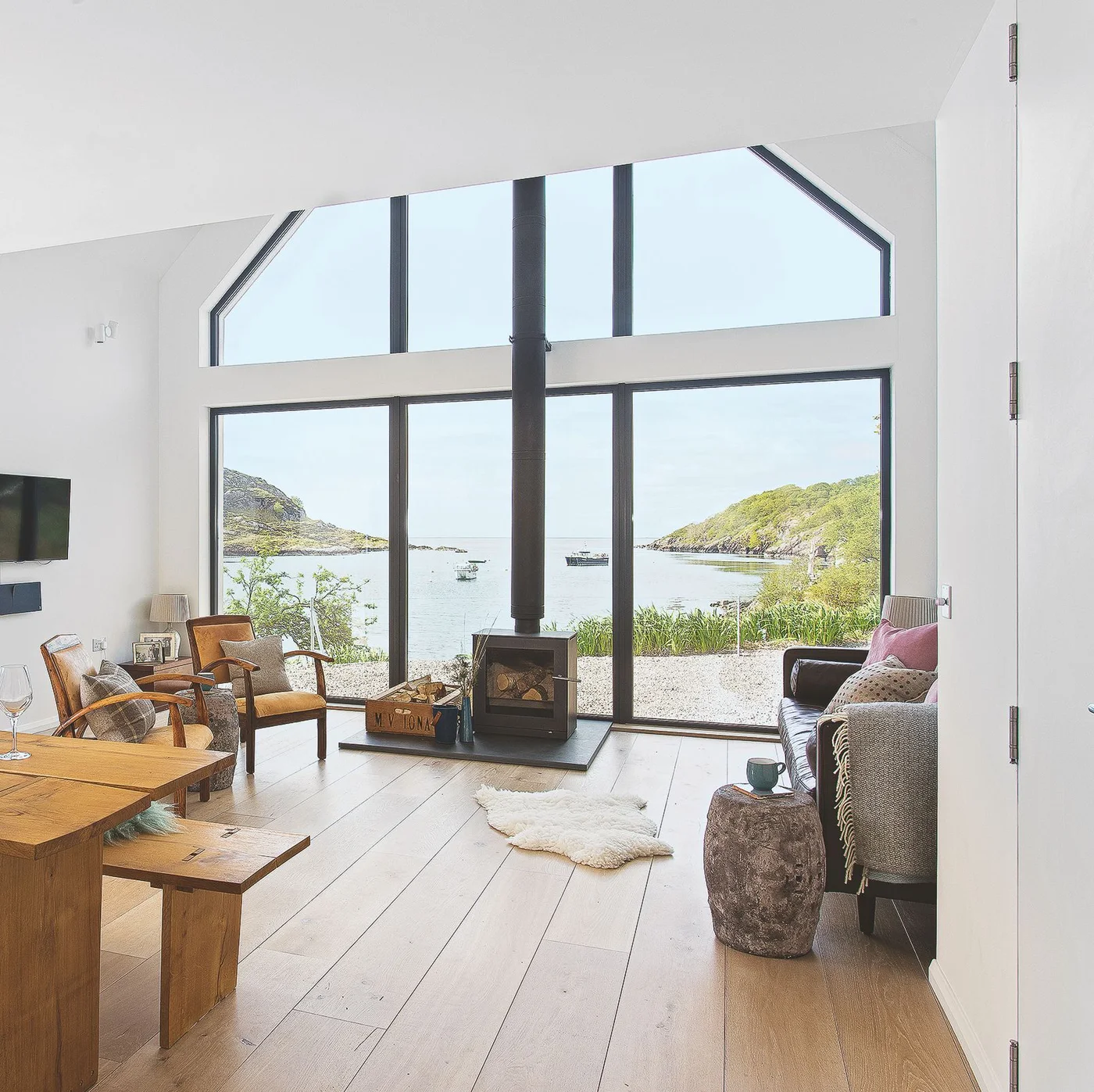 Wood armchairs and a dark sofa face a slim dark stove before broad windows above pale floorboards.