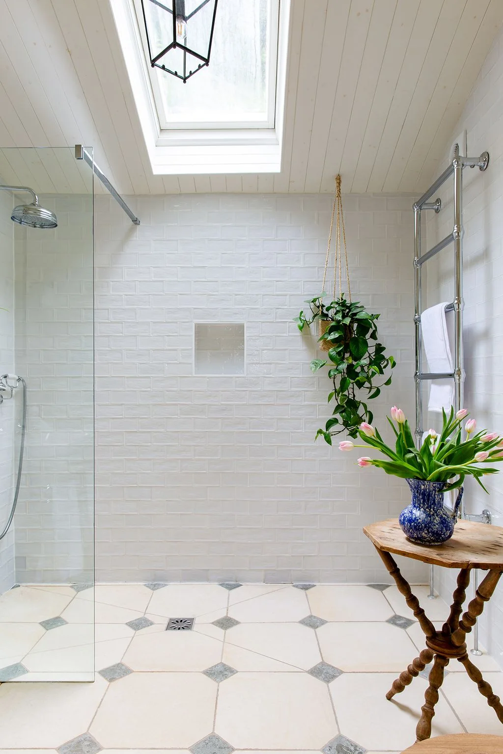 A clear shower screen stands beneath a skylight beside white wall tiles, pale octagonal floor tiles and a chrome towel rail.